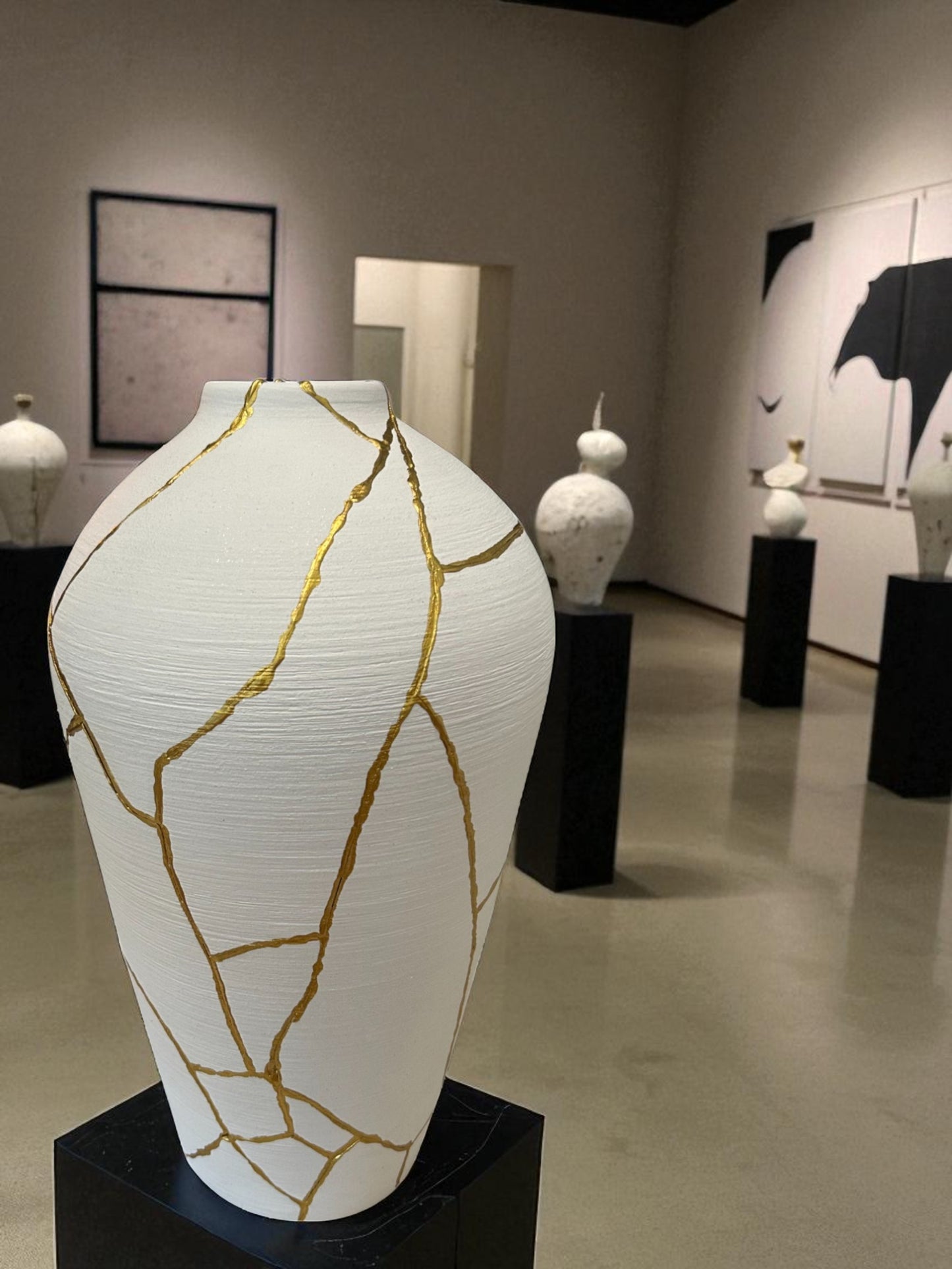 White ceramic vase with gold kintsugi repair lines displayed on black pedestal in modern art gallery setting