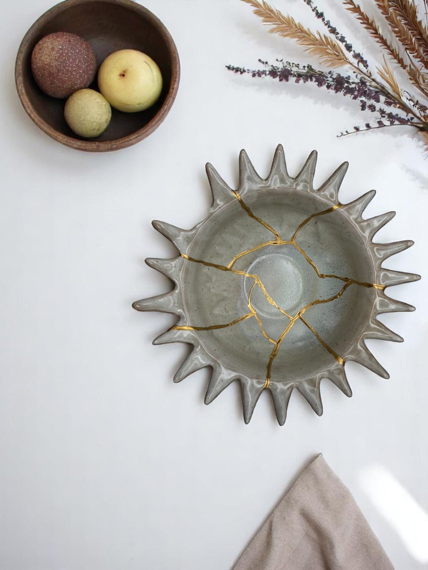 Gray ceramic bowl with sunburst edges repaired with gold kintsugi technique, styled with dried flowers and fruit bowl nearby