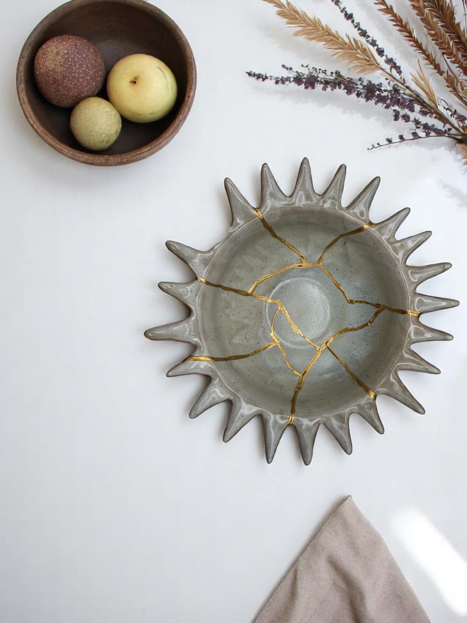 Gray ceramic bowl with sunburst edges repaired with gold kintsugi technique, styled with dried flowers and fruit bowl nearby