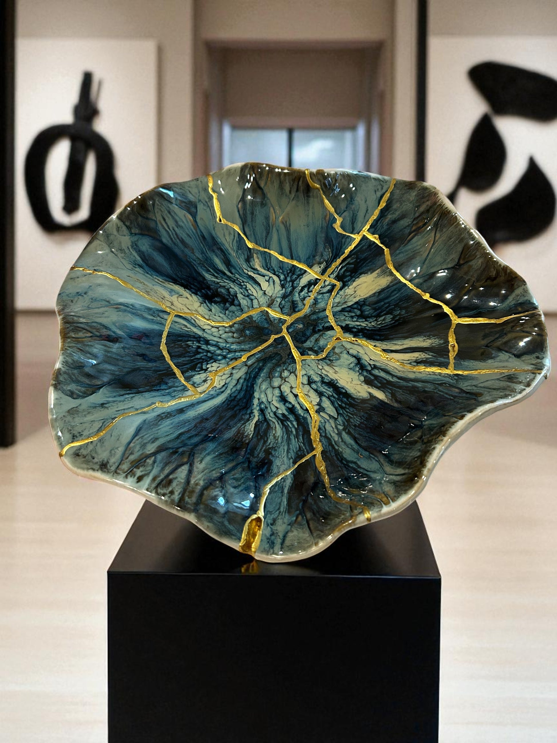 Blue ceramic bowl with gold repair lines in kintsugi style, displayed on black pedestal in modern gallery setting