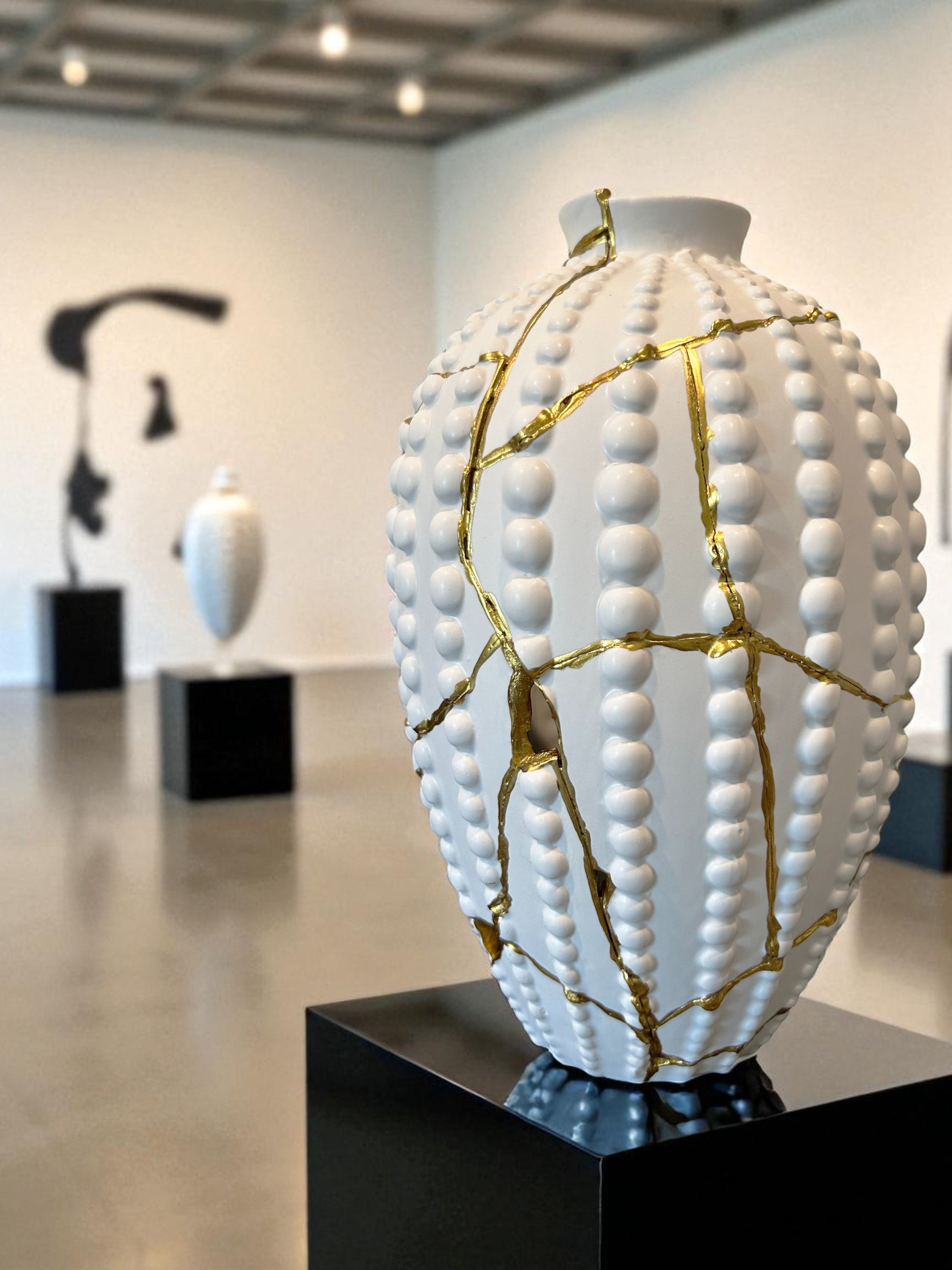 White ceramic vase with raised bead texture and gold repair lines, displayed on black pedestal in gallery setting