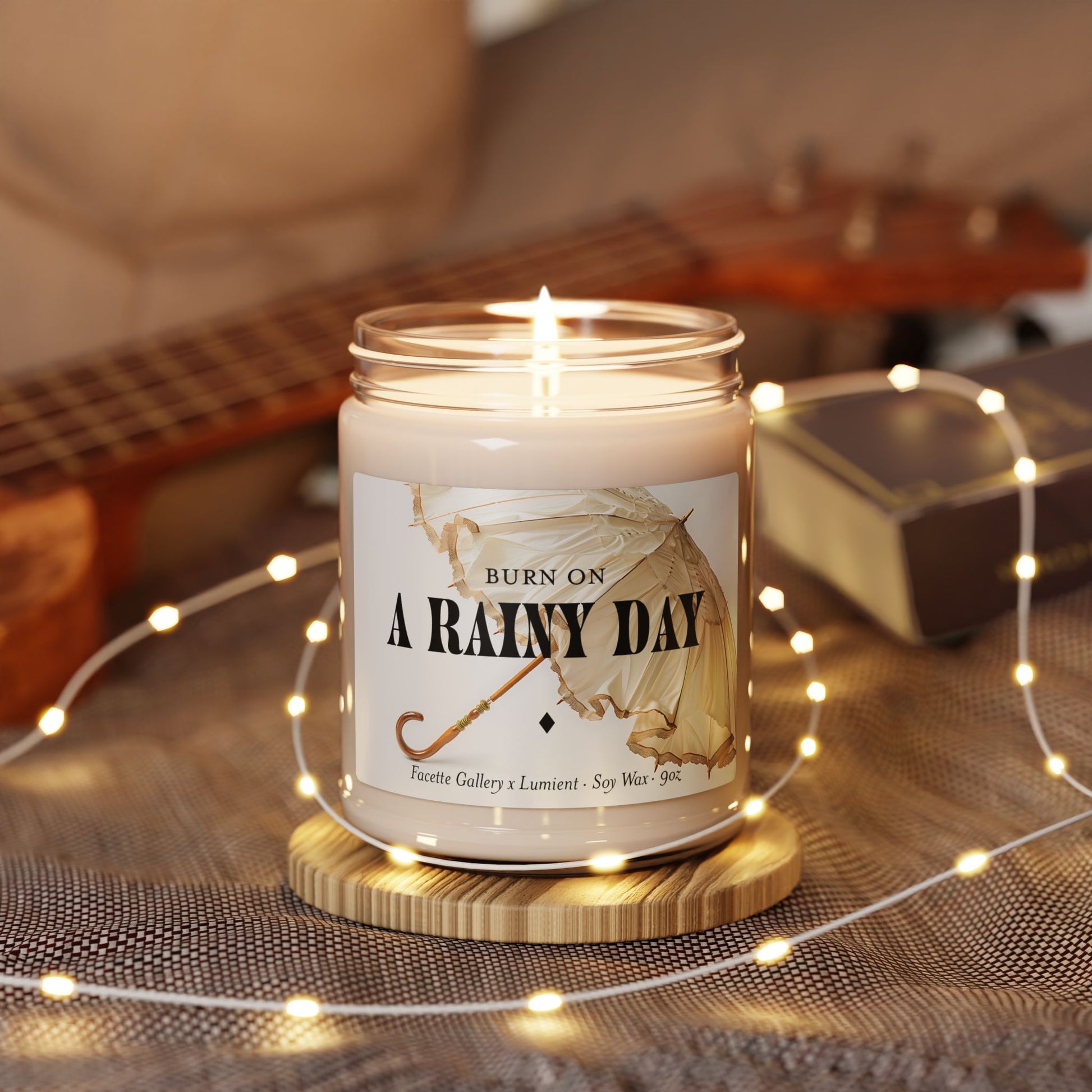 Lit 9oz soy candle in clear glass jar labeled "A Rainy Day" with umbrella design on wooden coaster and string lights