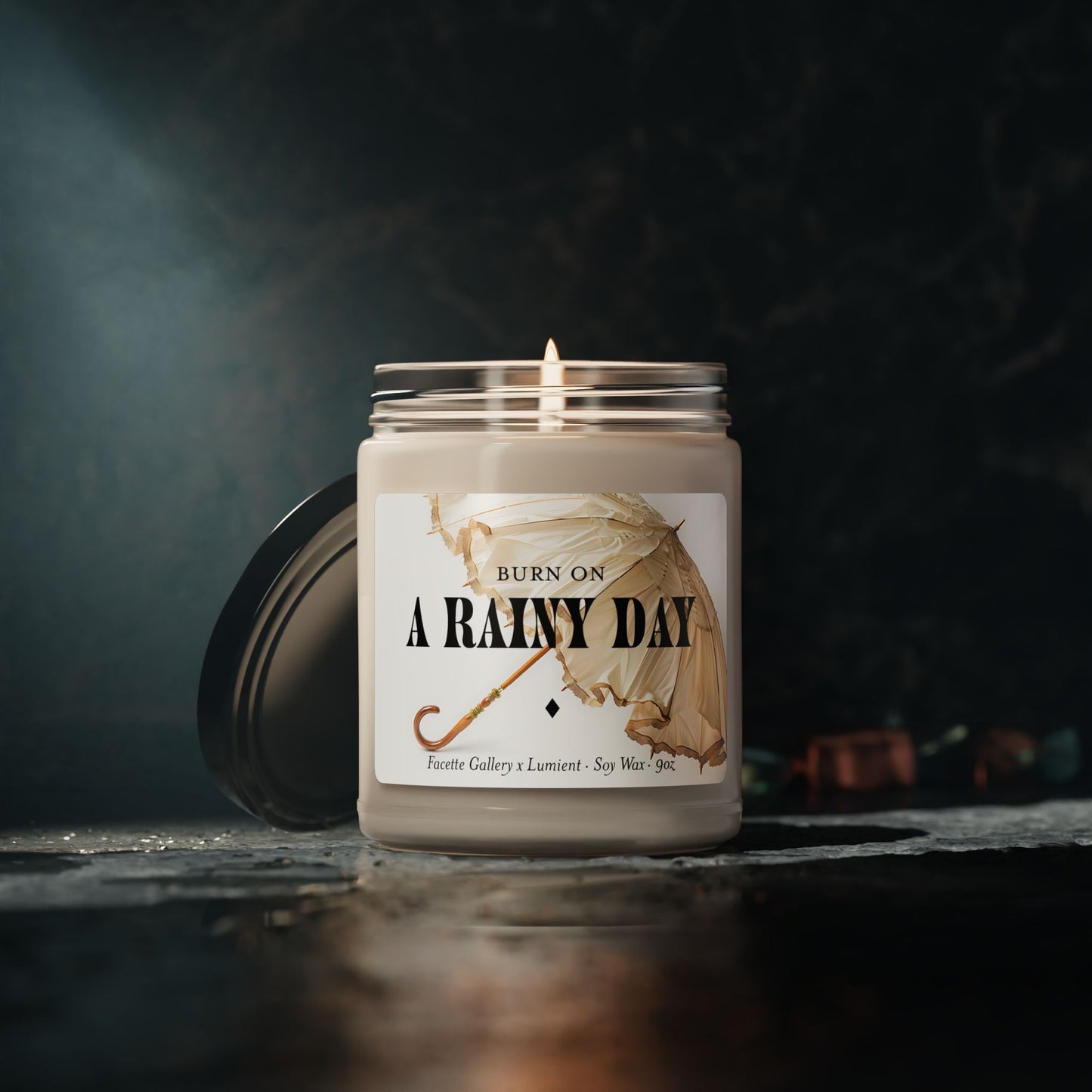 Lit "A Rainy Day" soy candle in clear glass jar with black lid on marble surface and dark moody background
