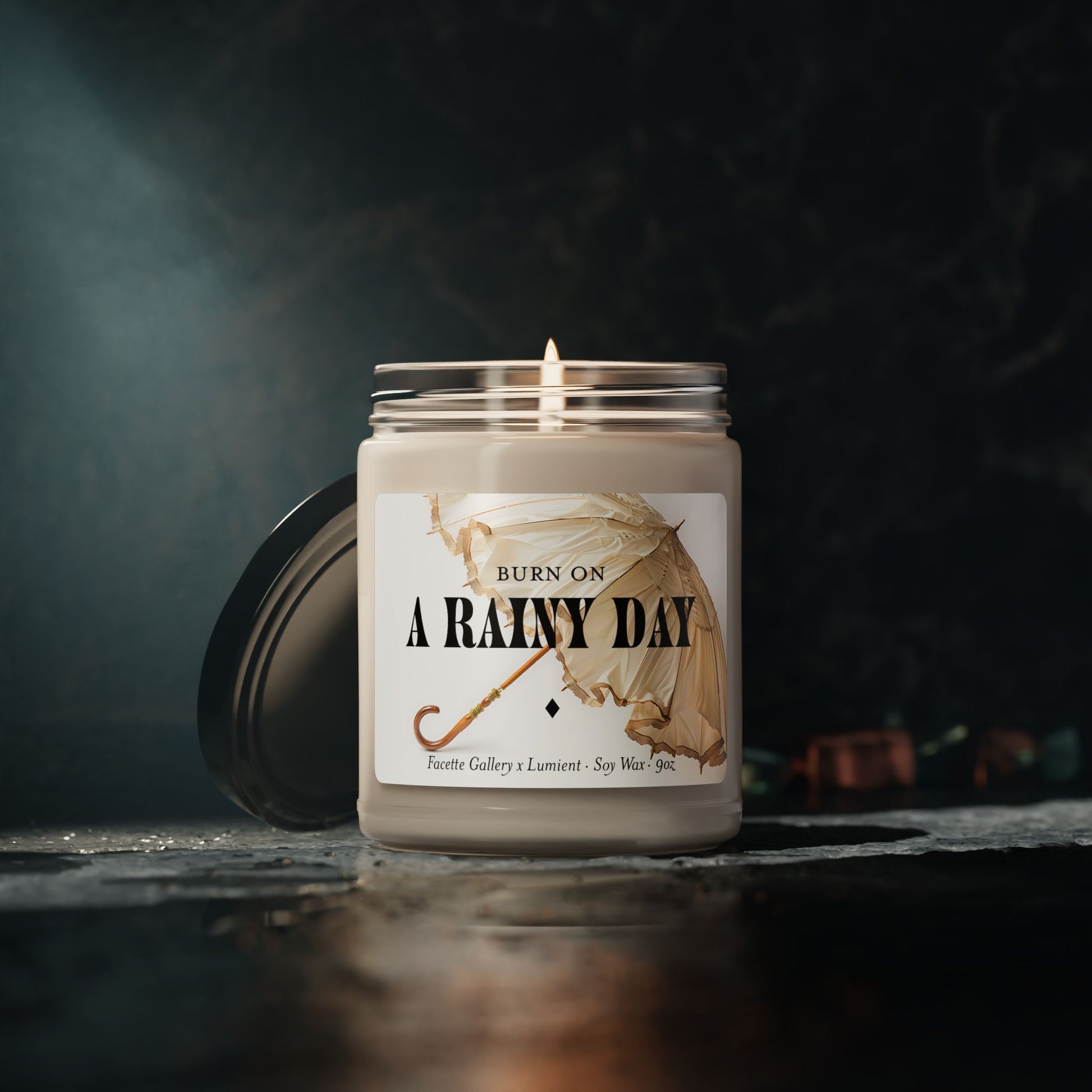 Lit "A Rainy Day" soy candle in clear glass jar with black lid on marble surface and dark moody background