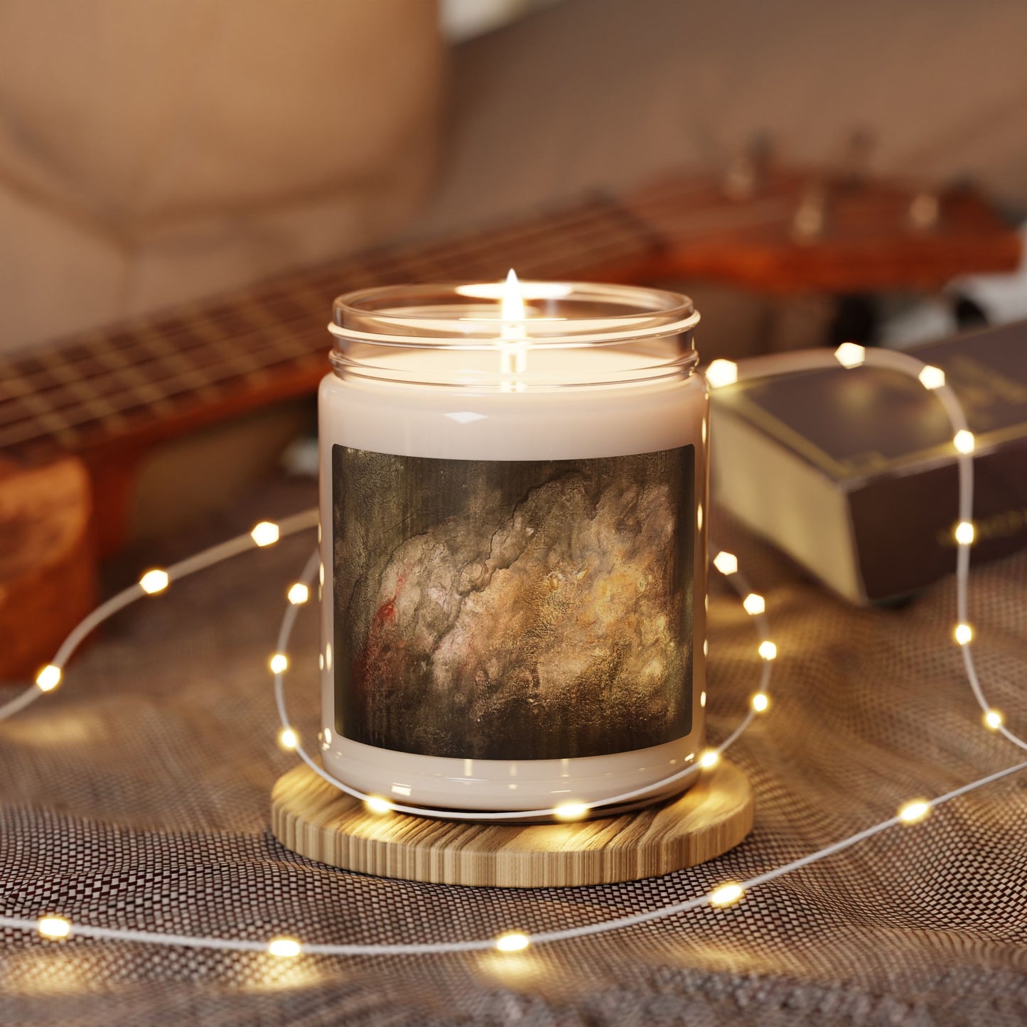 Lit 9oz soy candle in glass jar featuring abstract painting label, surrounded by warm fairy lights and guitar background