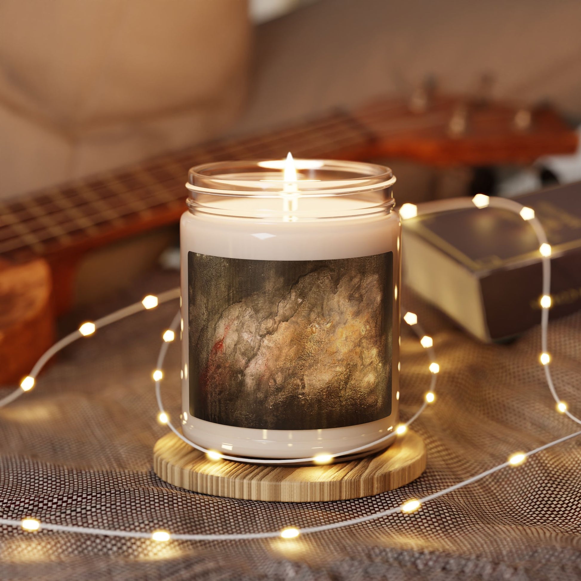 Lit 9oz soy candle in glass jar featuring abstract painting label, surrounded by warm fairy lights and guitar background