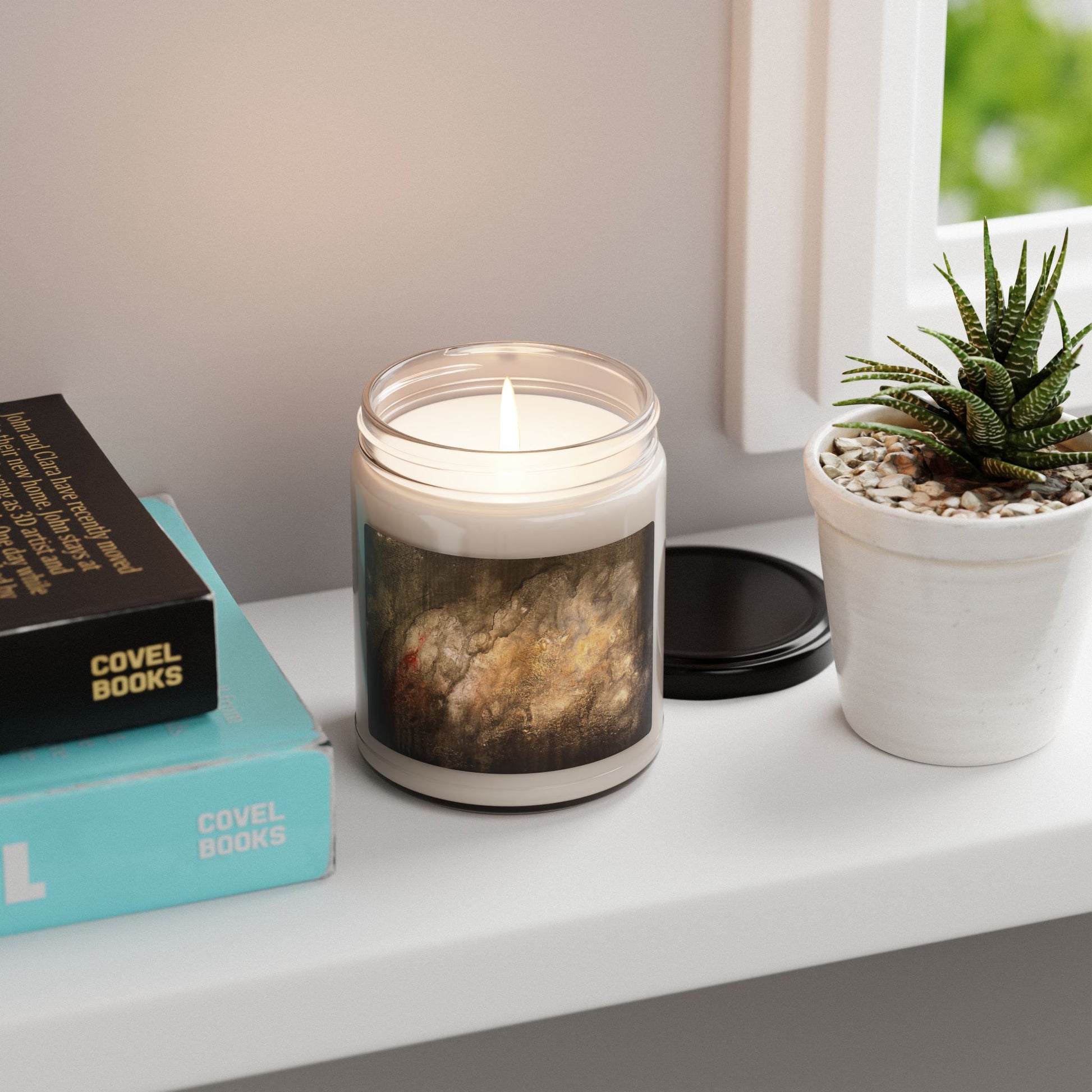 Lit 9oz soy candle in clear glass jar with abstract painting by Ben Woolfitt, next to books and potted succulent on windowsill