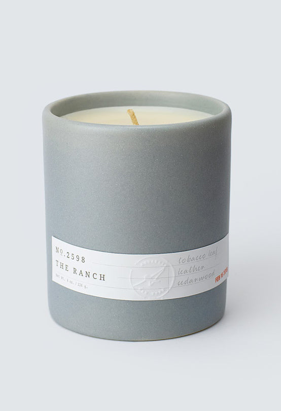 Gray ceramic candle labeled Aerangis No. 2598 The Ranch with tobacco, leather, and cedarwood scent notes on white background