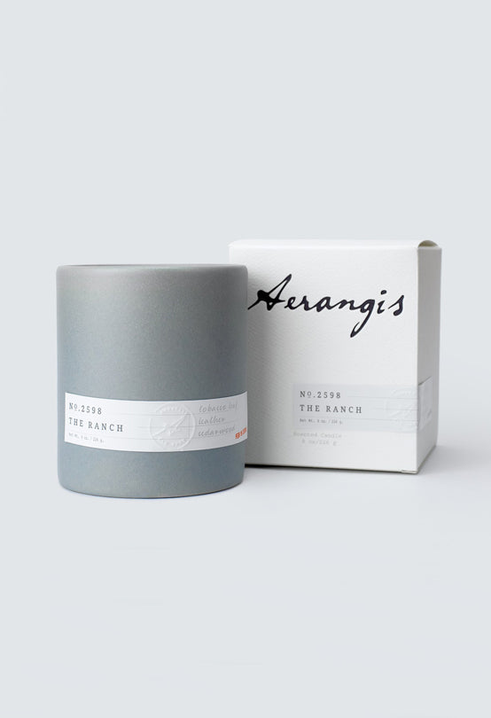 Gray cylindrical candle container labeled Aerangis No. 2598 The Ranch next to white packaging box on light background