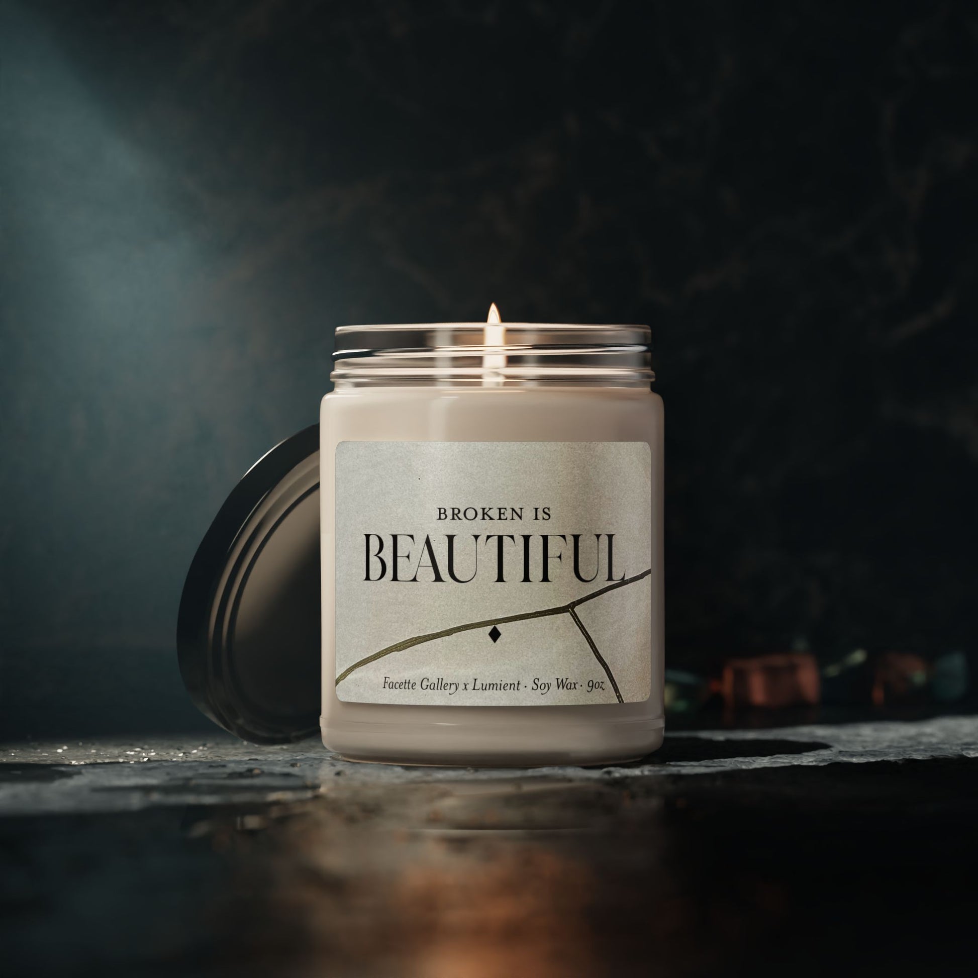 Lit beige soy candle in glass jar with "Broken is Beautiful" label and black lid on dark textured surface