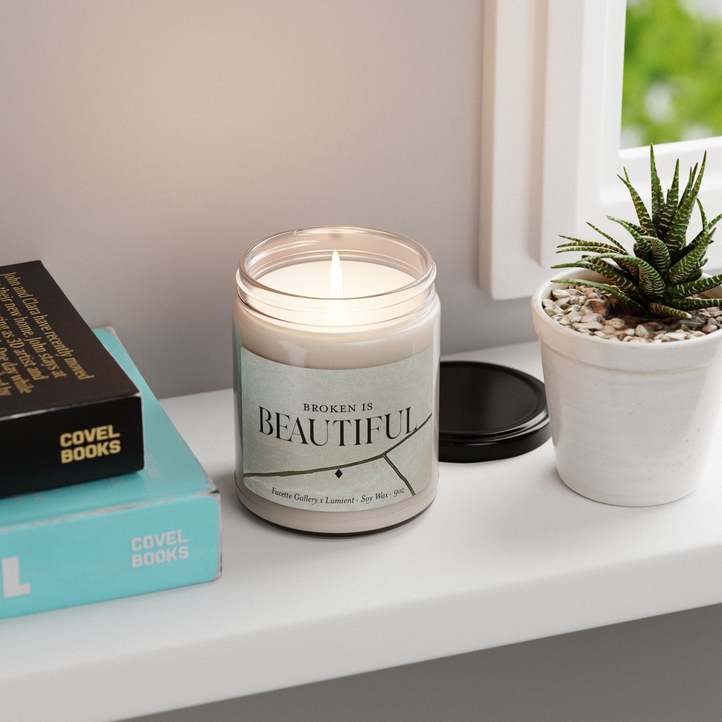 Lit "Broken Is Beautiful" soy candle in clear glass jar with beige label, placed on white windowsill next to books and small plant