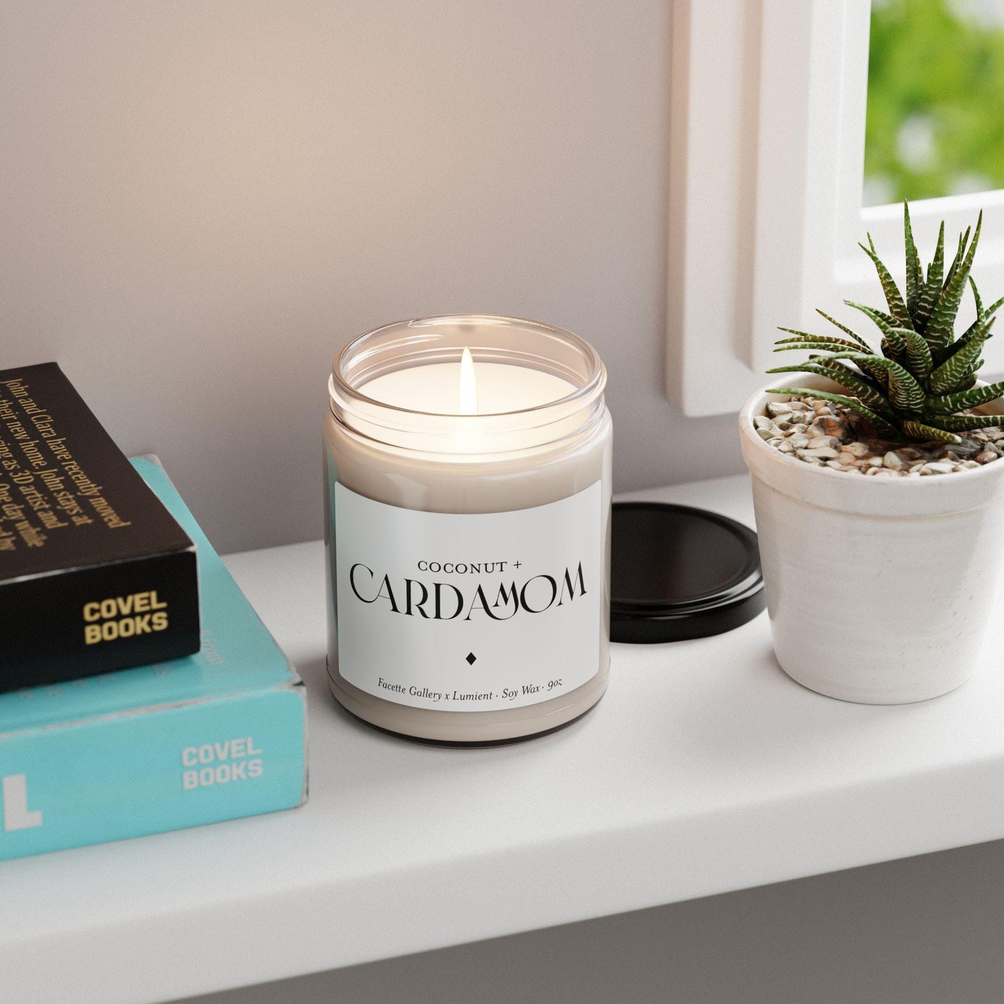 Lit Coconut + Cardamom soy candle in 9oz clear glass jar with black and white label on a white windowsill next to plant and books