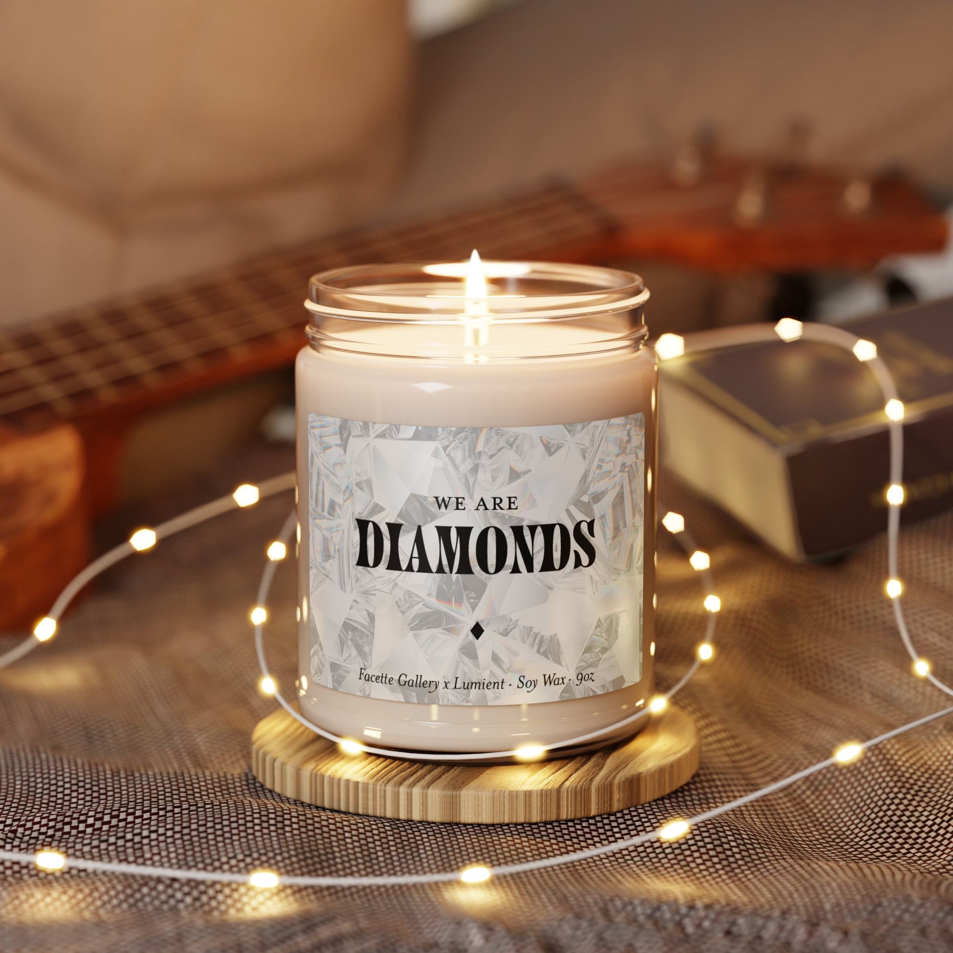 Facette Gallery "We Are Diamonds" soy candle in 9oz glass jar with lit wick, surrounded by string lights on fabric.