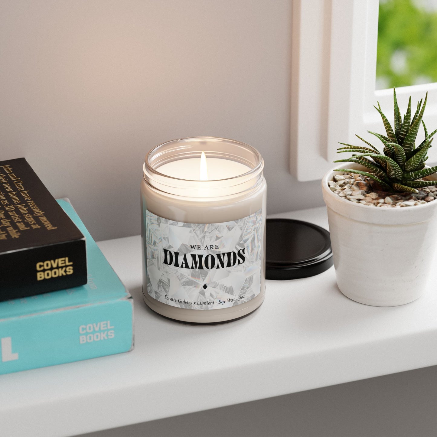 Facette Gallery "We Are Diamonds" soy candle in 9oz glass jar with lit wick, next to books and succulent on white shelf