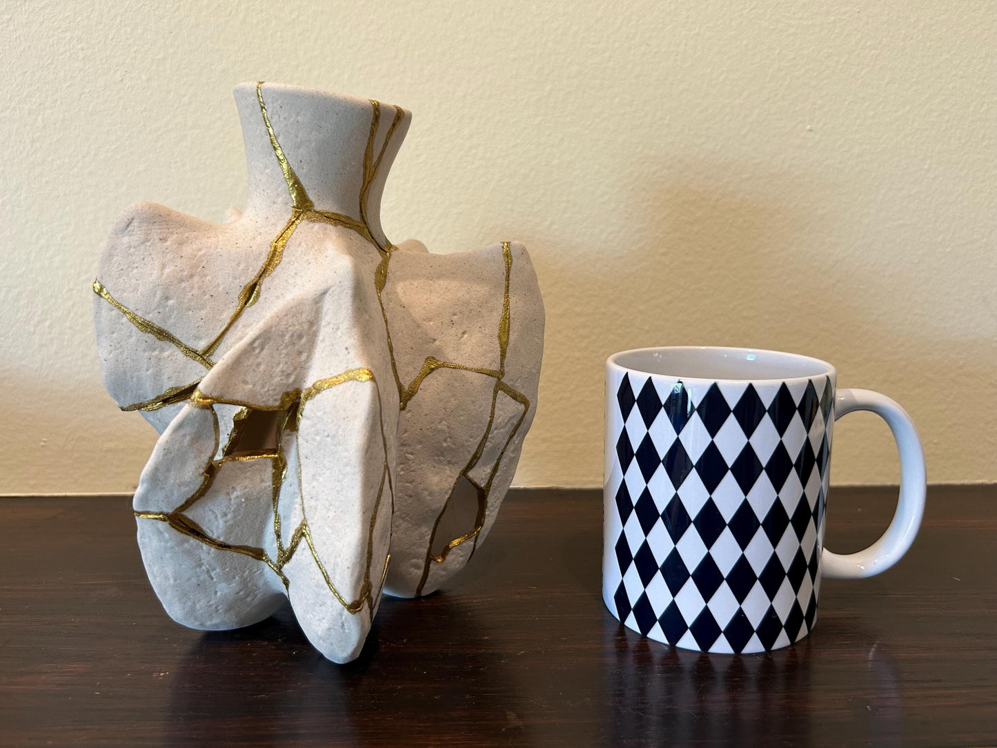 Modern Kintsugi fossil vase with hand-painted gold repair lines next to a white mug with black diamond pattern on wooden surface