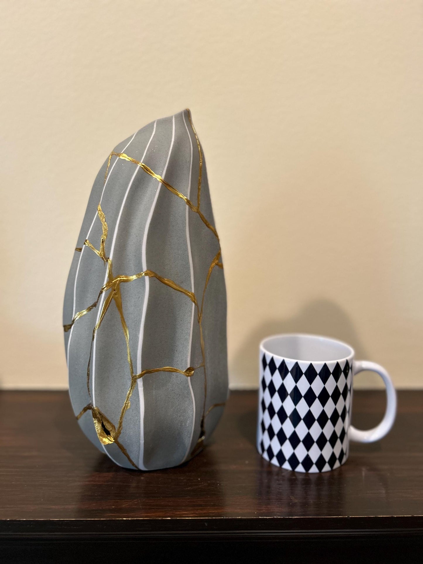 Modern Kintsugi melon-shaped vase with gold repair lines and white stripes beside black and white diamond patterned mug on wood table