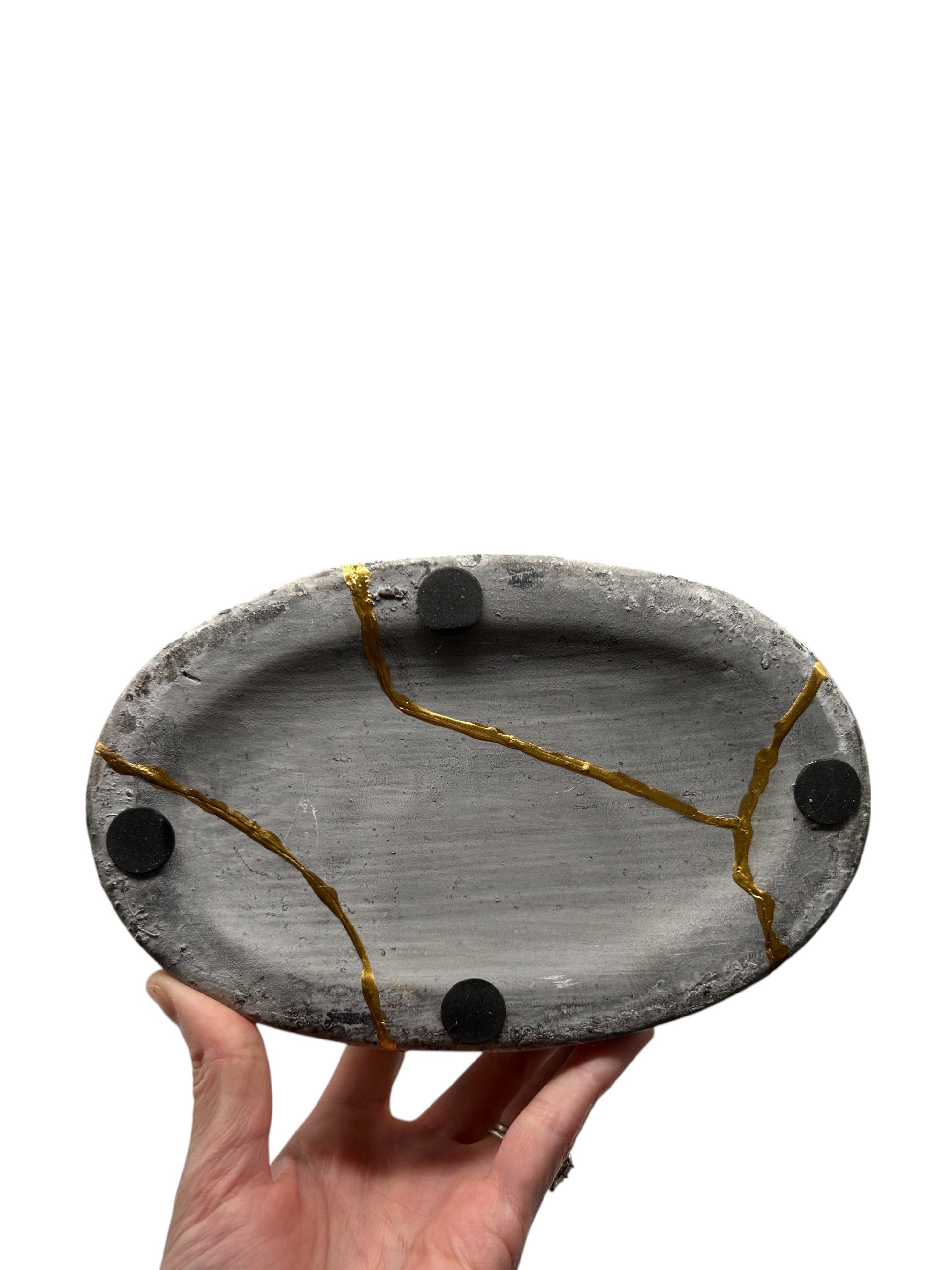 Bottom view of oval gray ceramic vase with hand-painted gold kintsugi cracks and black felt pads held by hand against white background