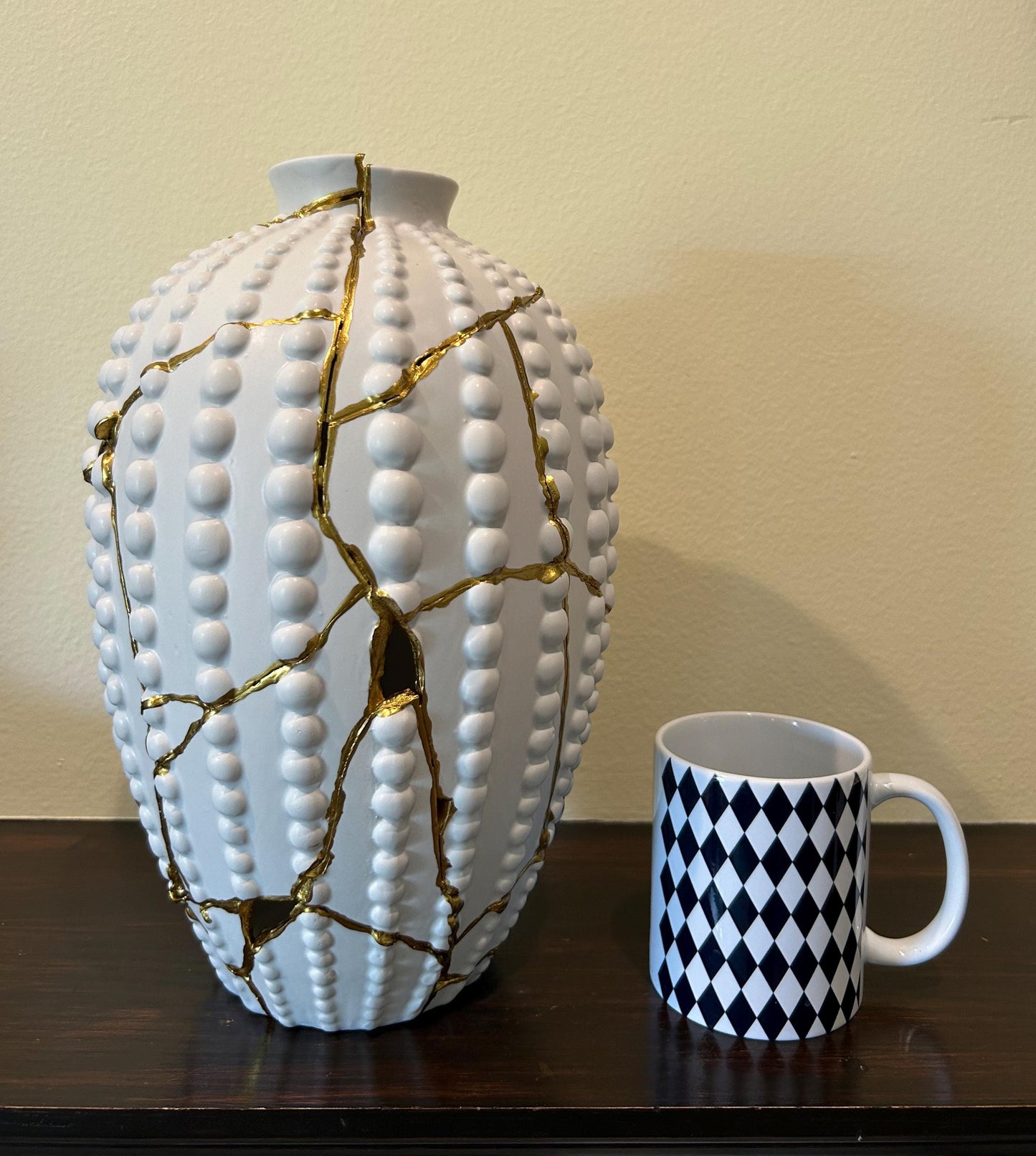 White modern kintsugi sea urchin vase with gold repaired cracks, next to white mug with black diamond pattern on wooden surface