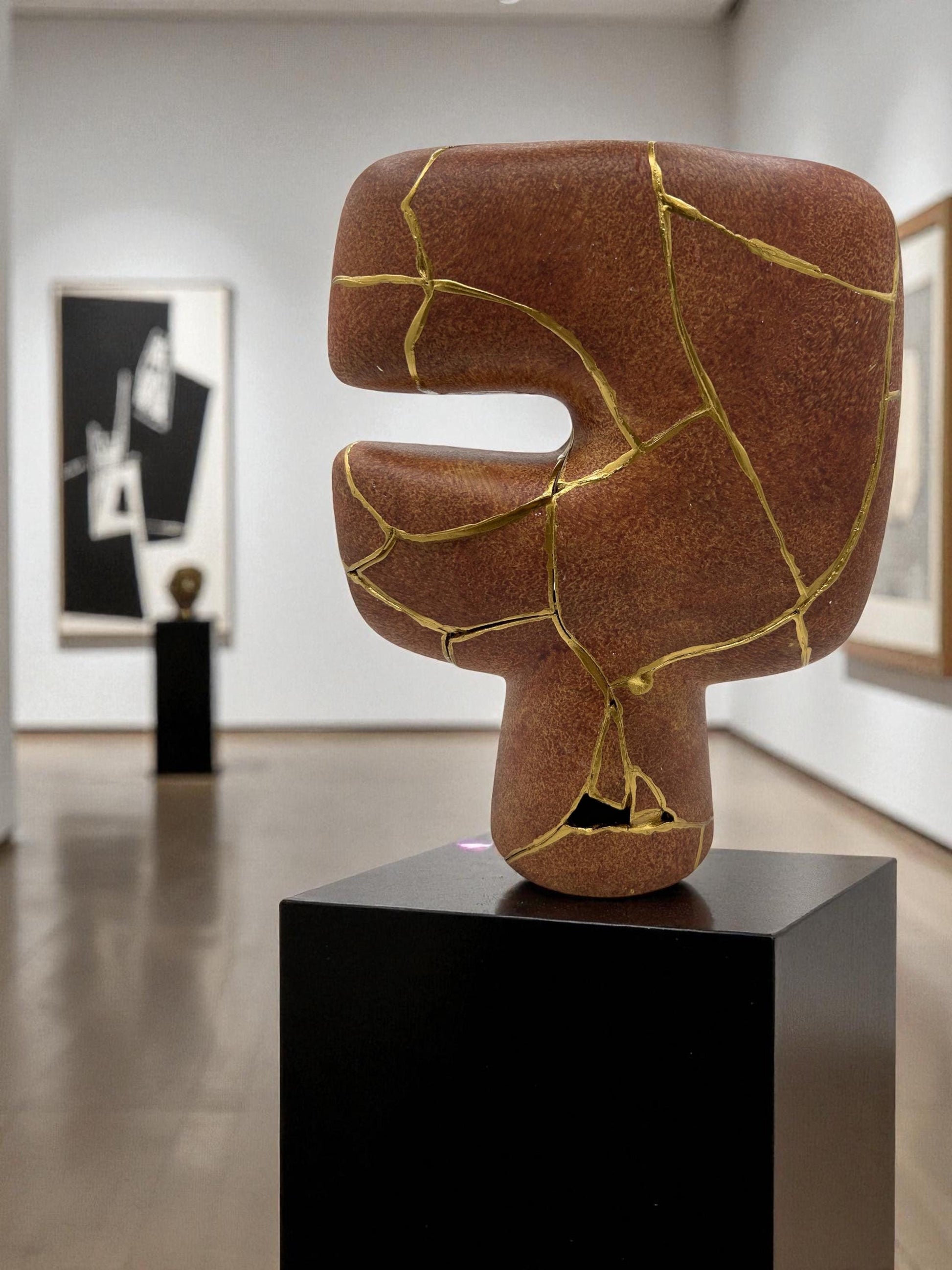 Abstract brown kintsugi vase with gold repair lines displayed on black pedestal in a modern gallery setting