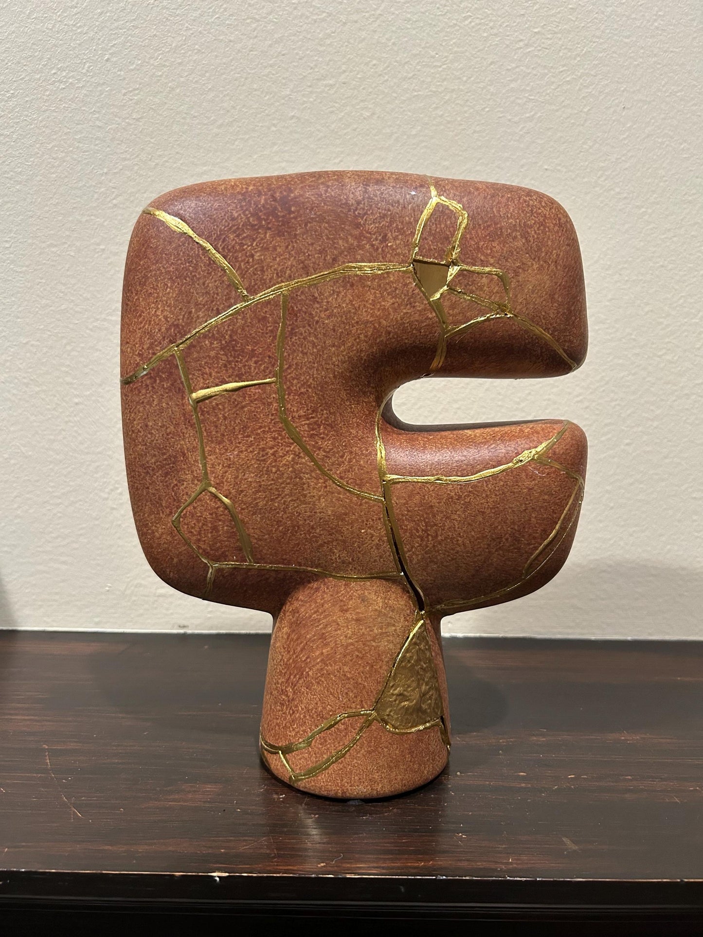 Abstract brown kintsugi vase with gold repair lines, modern tribal shape, 10in tall, on dark wood surface and beige wall background