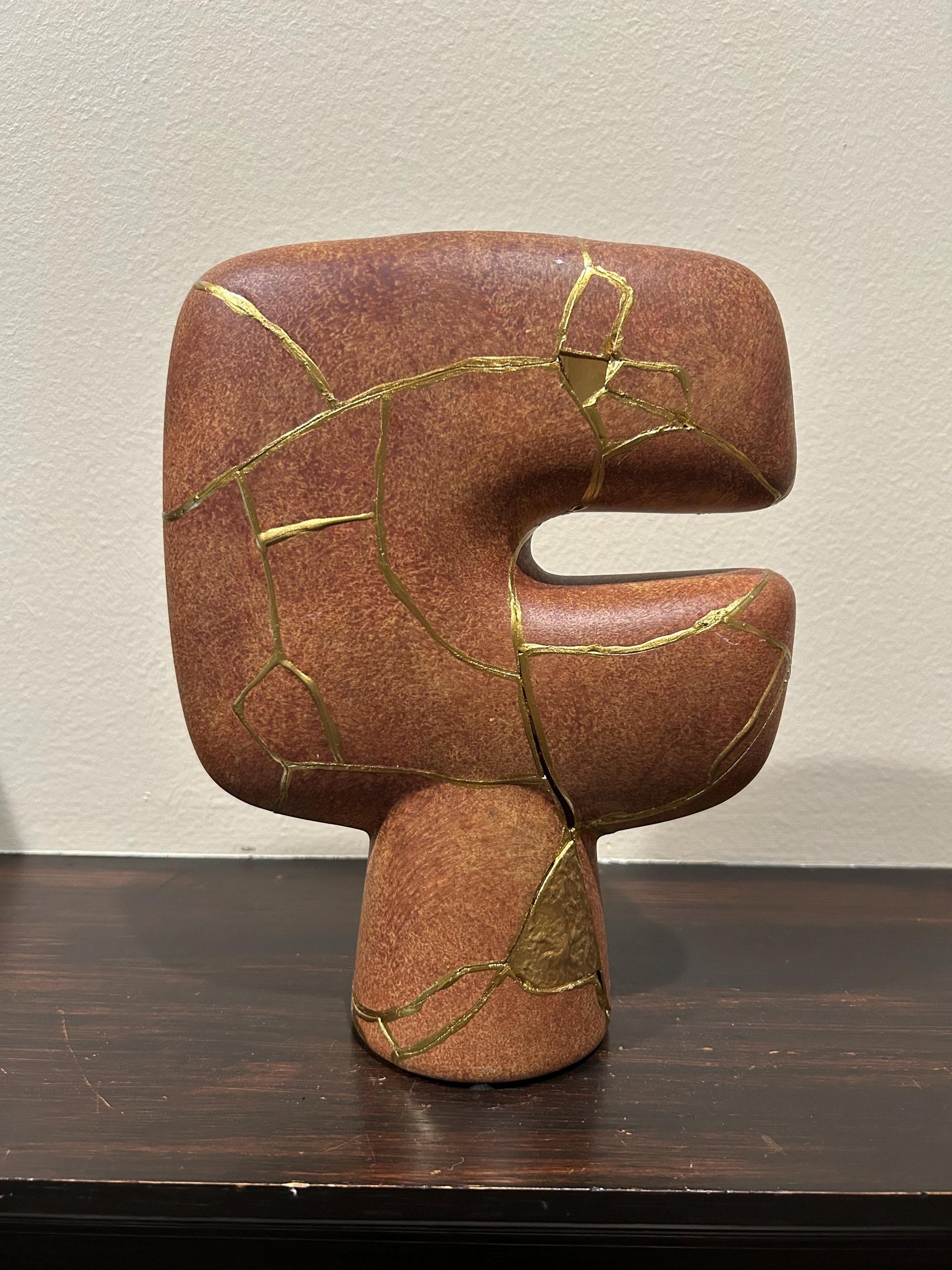 Abstract brown kintsugi vase with gold repair lines, modern tribal shape, 10in tall, on dark wood surface and beige wall background