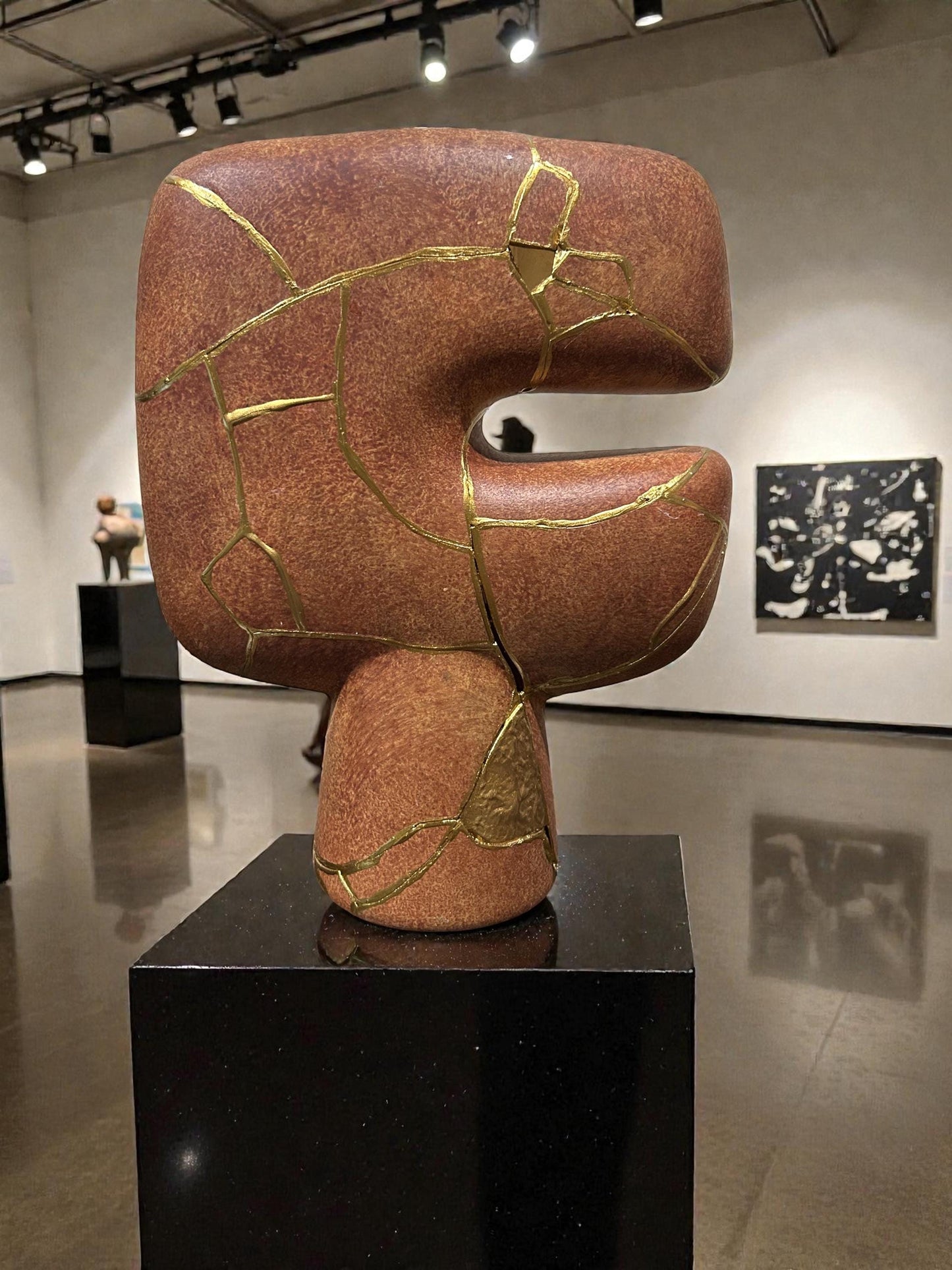 Abstract modern kintsugi ceramic vase with gold repair lines, earthy brown finish, displayed on black pedestal in gallery space