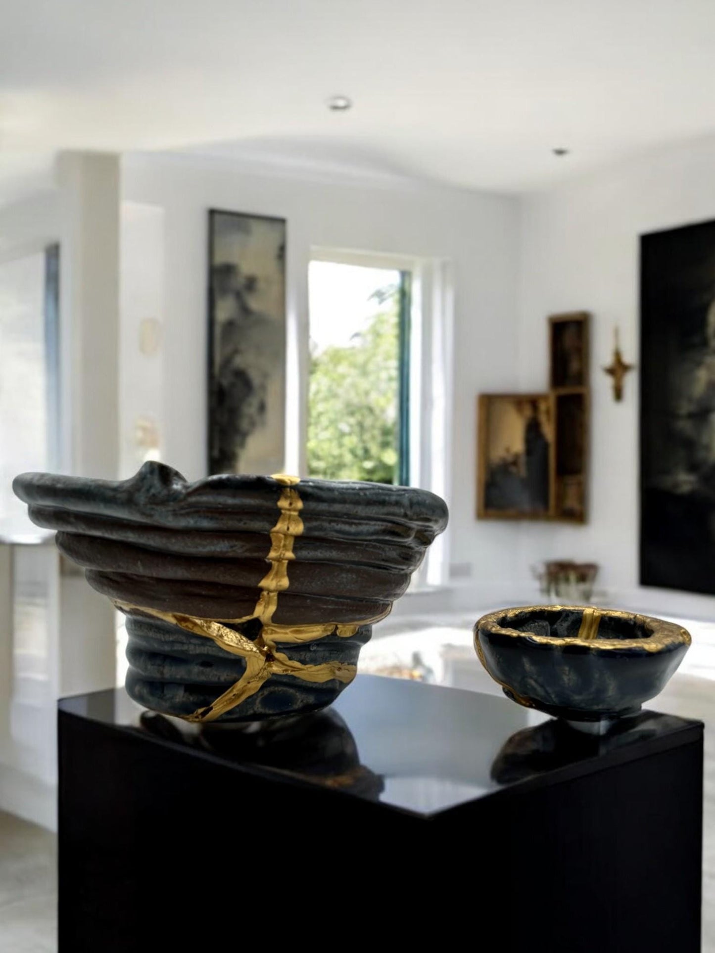 Two-piece set of modern Kintsugi ceramic bowls with hand-painted gold repairs on black display pedestal in bright room