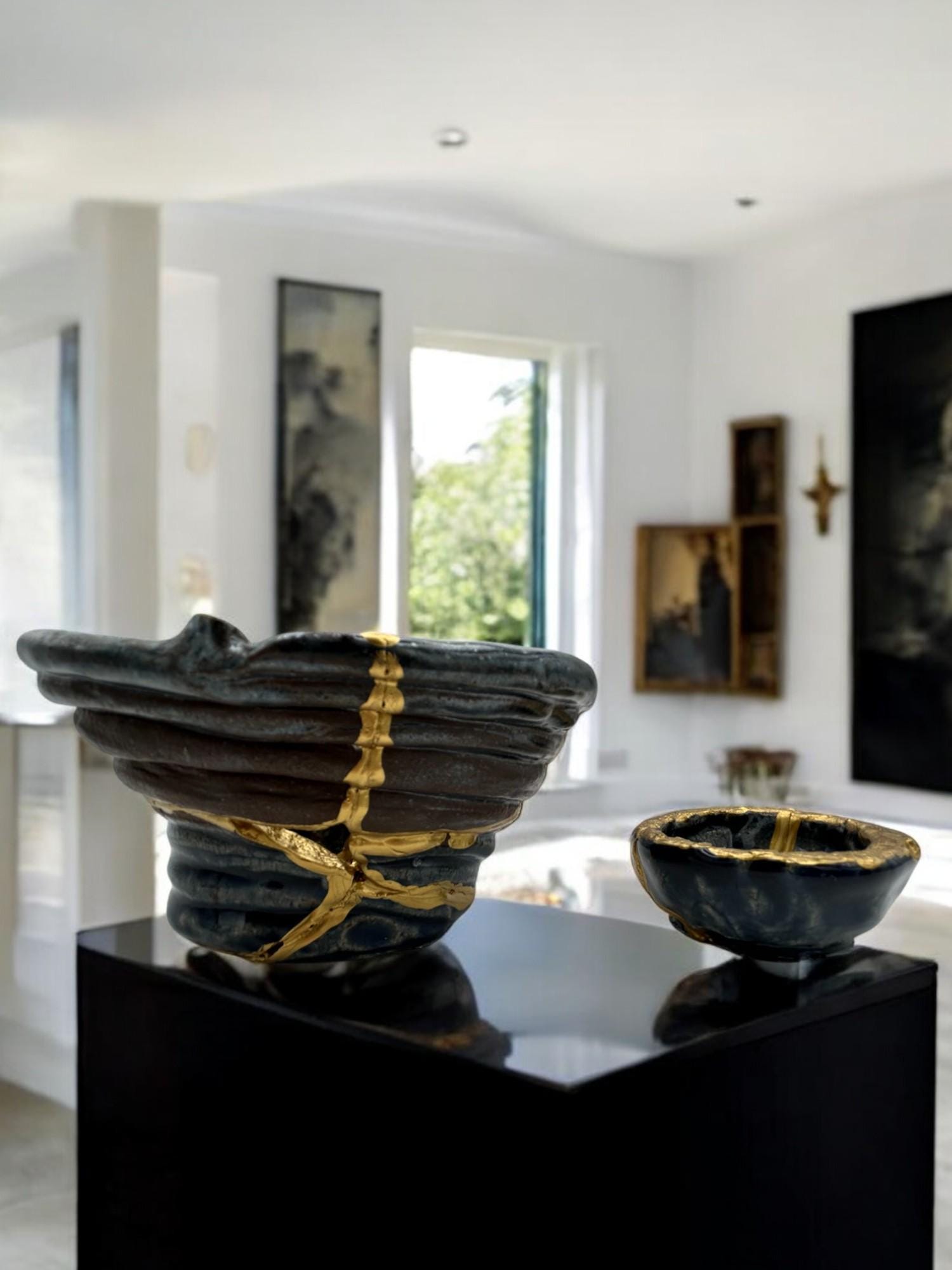 Two-piece set of modern Kintsugi ceramic bowls with hand-painted gold repairs on black display pedestal in bright room
