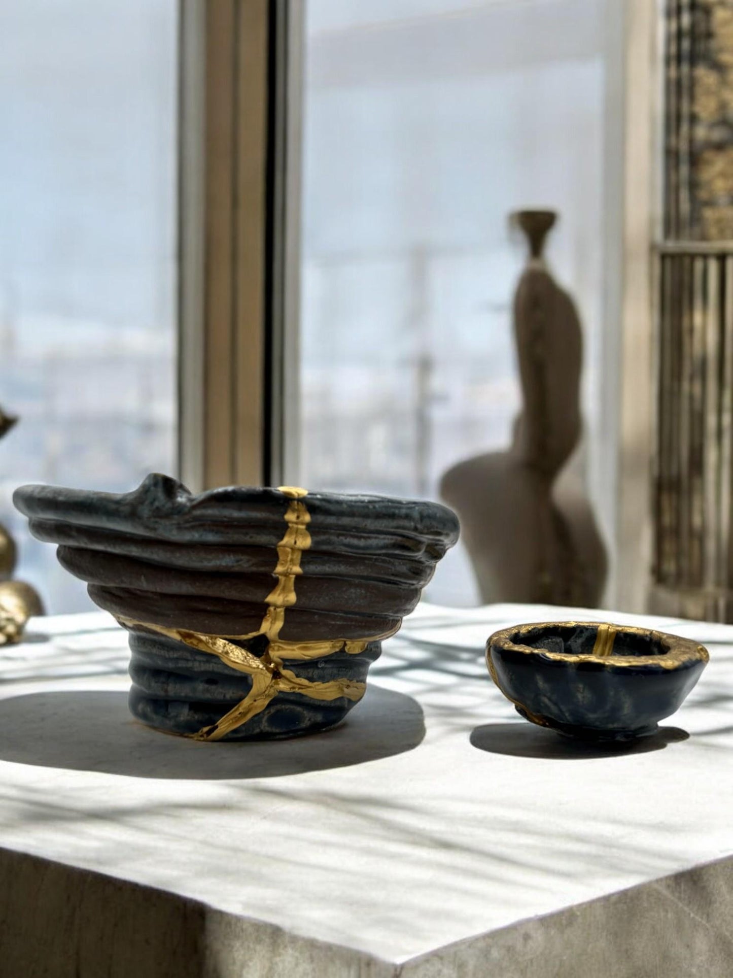 Two-piece set of modern black kintsugi bowls with hand-painted gold repairs, displayed on a sunlit stone surface.