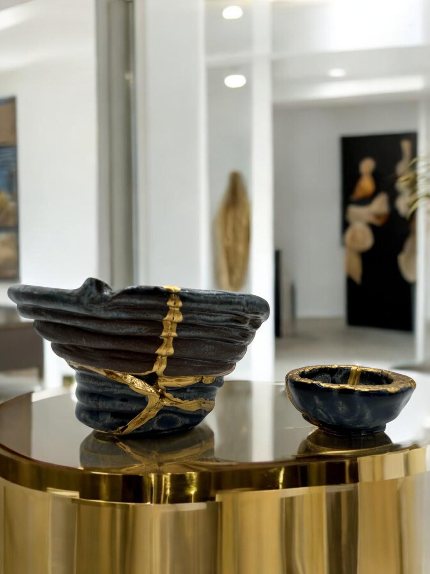 Two-piece set of modern kintsugi bowls in dark ceramic with hand-painted gold repairs on a reflective gold surface