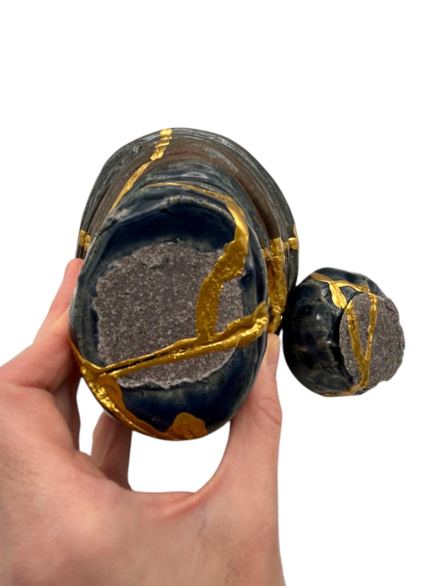 Two-piece modern kintsugi pottery bowls in dark blue with hand-painted gold repair lines, held in hand on white background