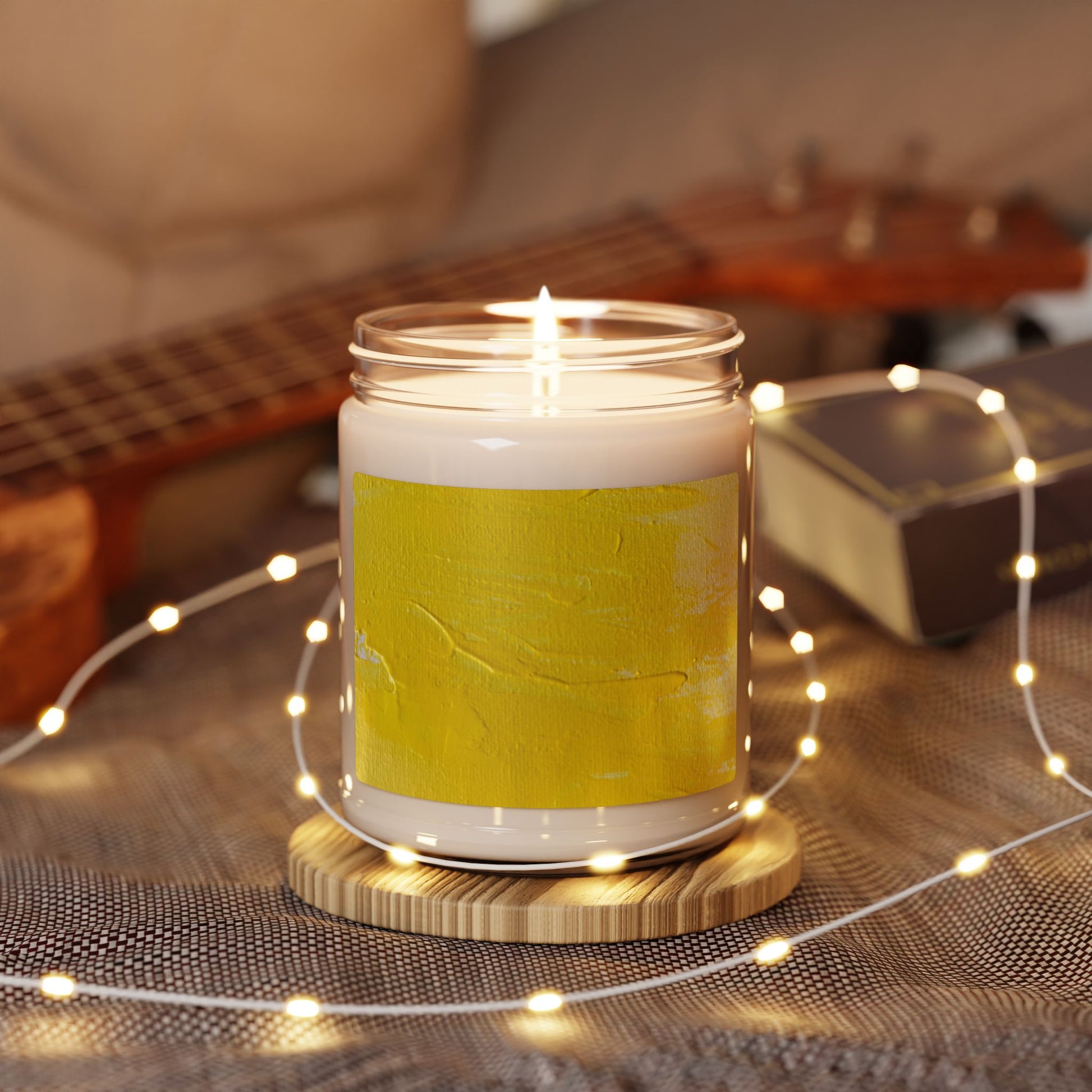 9oz soy candle in glass jar with yellow abstract painting label, lit and surrounded by string lights on wooden coaster