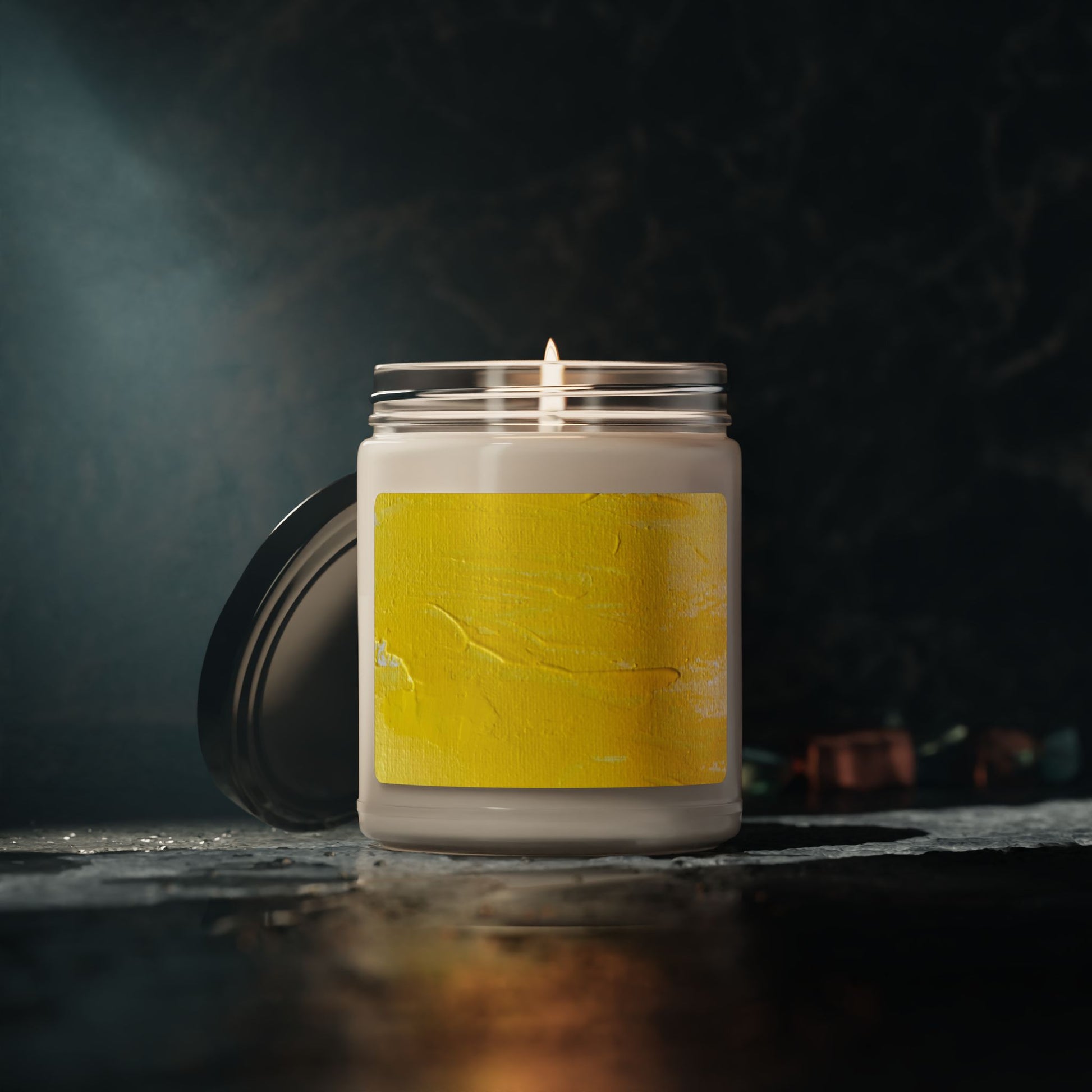 Lit 9oz soy candle in glass jar with yellow abstract painting label and black lid on dark surface