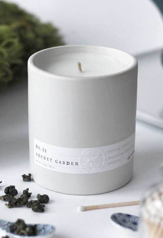 White ceramic candle labeled No. 51 Secret Garden with natural wax and loose tea leaves on white surface