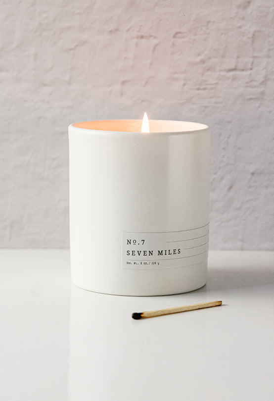 Lit white ceramic candle labeled No. 7 Seven Miles with a burnt matchstick on white surface and textured background