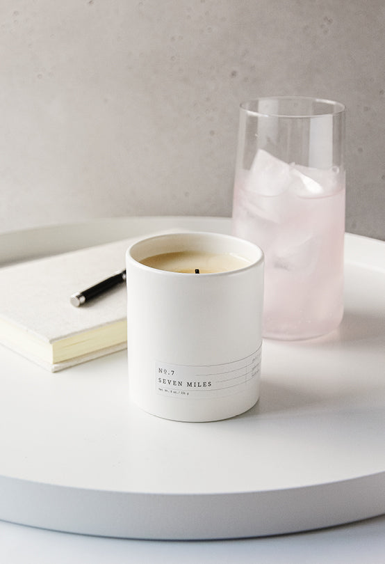 White ceramic candle labeled No. 7 Seven Miles on a round white tray with notebook, pen, and iced pink drink nearby