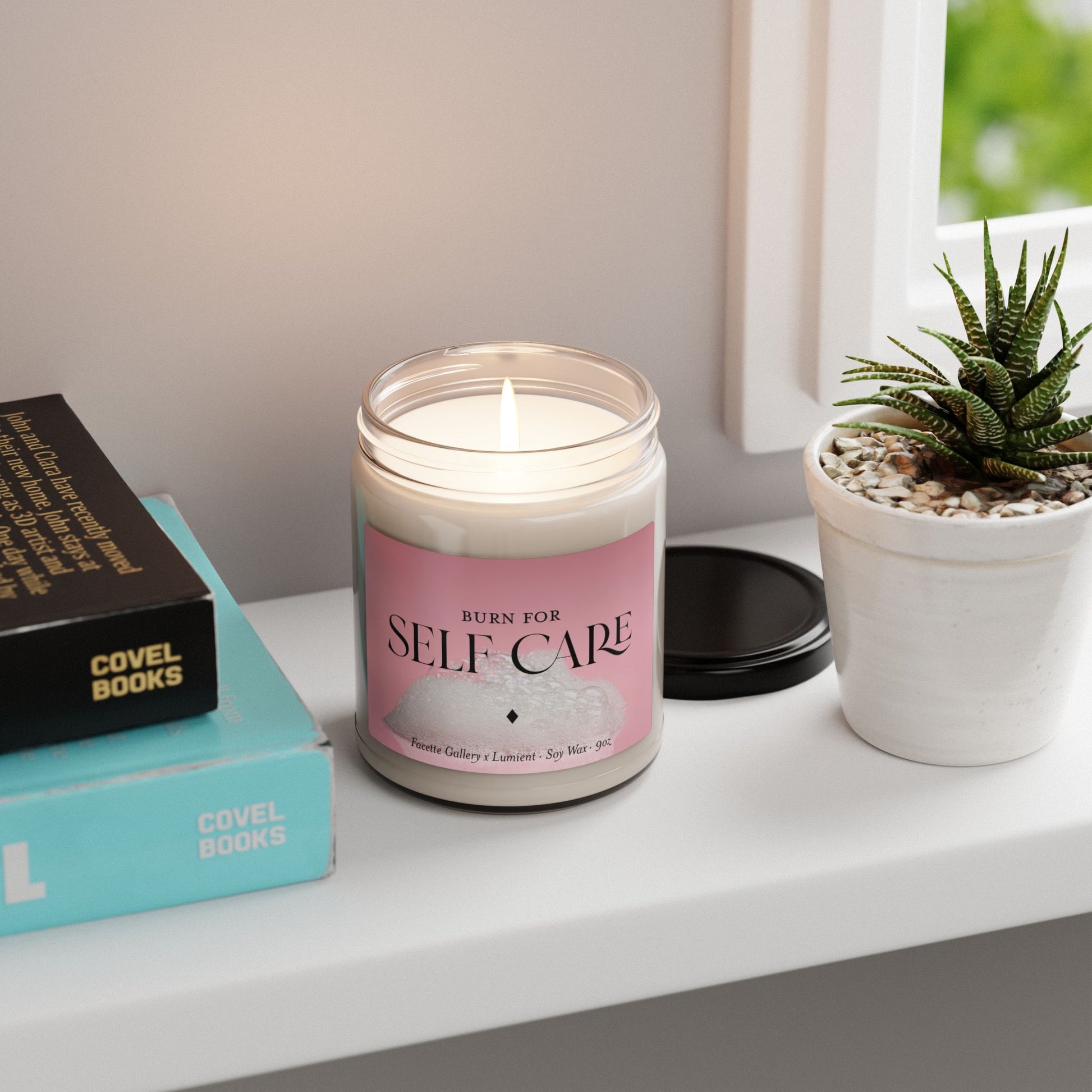 Lit self care soy candle in 9oz glass jar with pink label, placed on white windowsill next to books and succulent plant