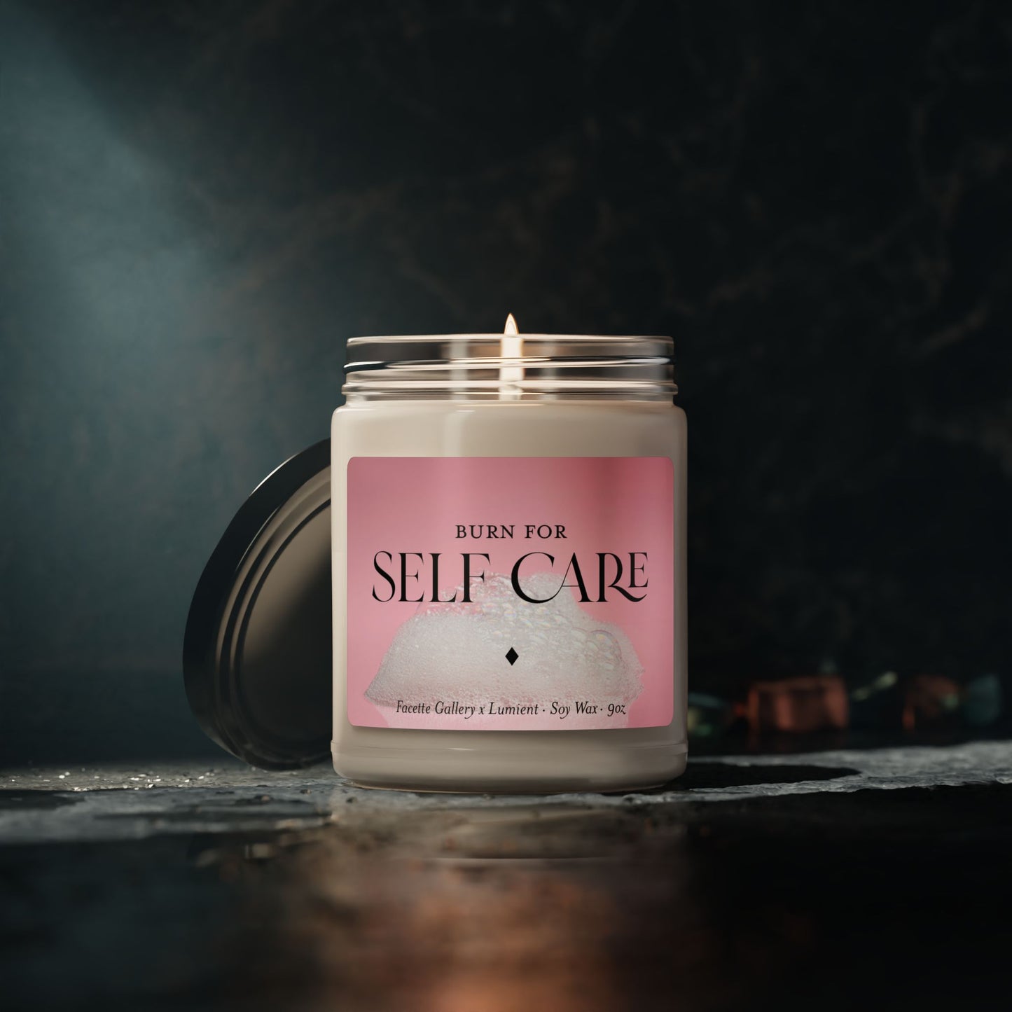9oz soy wax self care candle in glass jar with pink bubble bath label and lit cotton wick on dark background