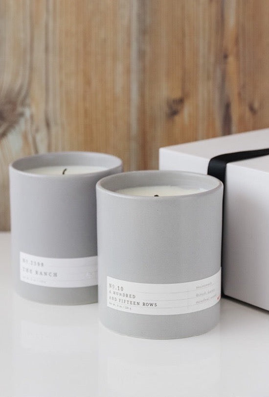 Two gray ceramic candles with minimal white labels beside a white gift box with black ribbon on wood background