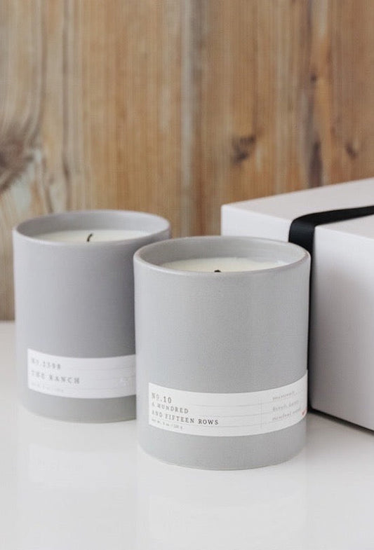 Two gray ceramic candles with minimal white labels beside a white gift box with black ribbon on wood background