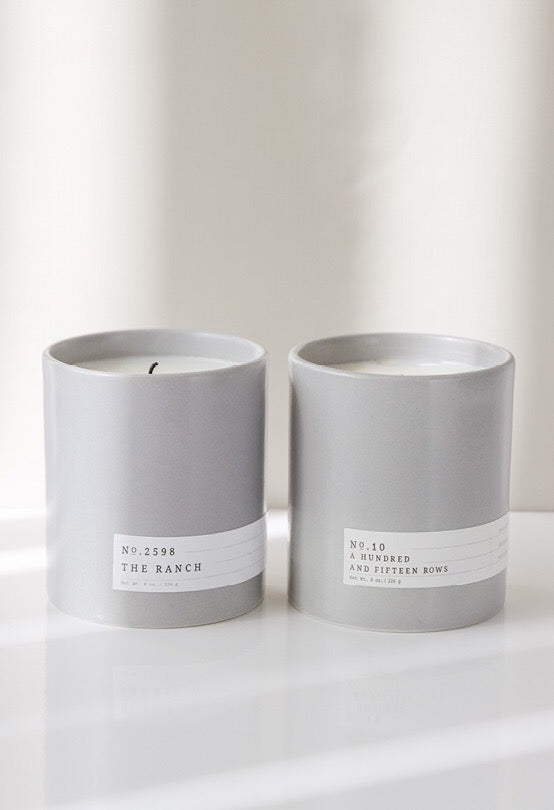 Two gray ceramic candles with minimalist white labels reading The Ranch and A Hundred and Fifteen Rows on white surface