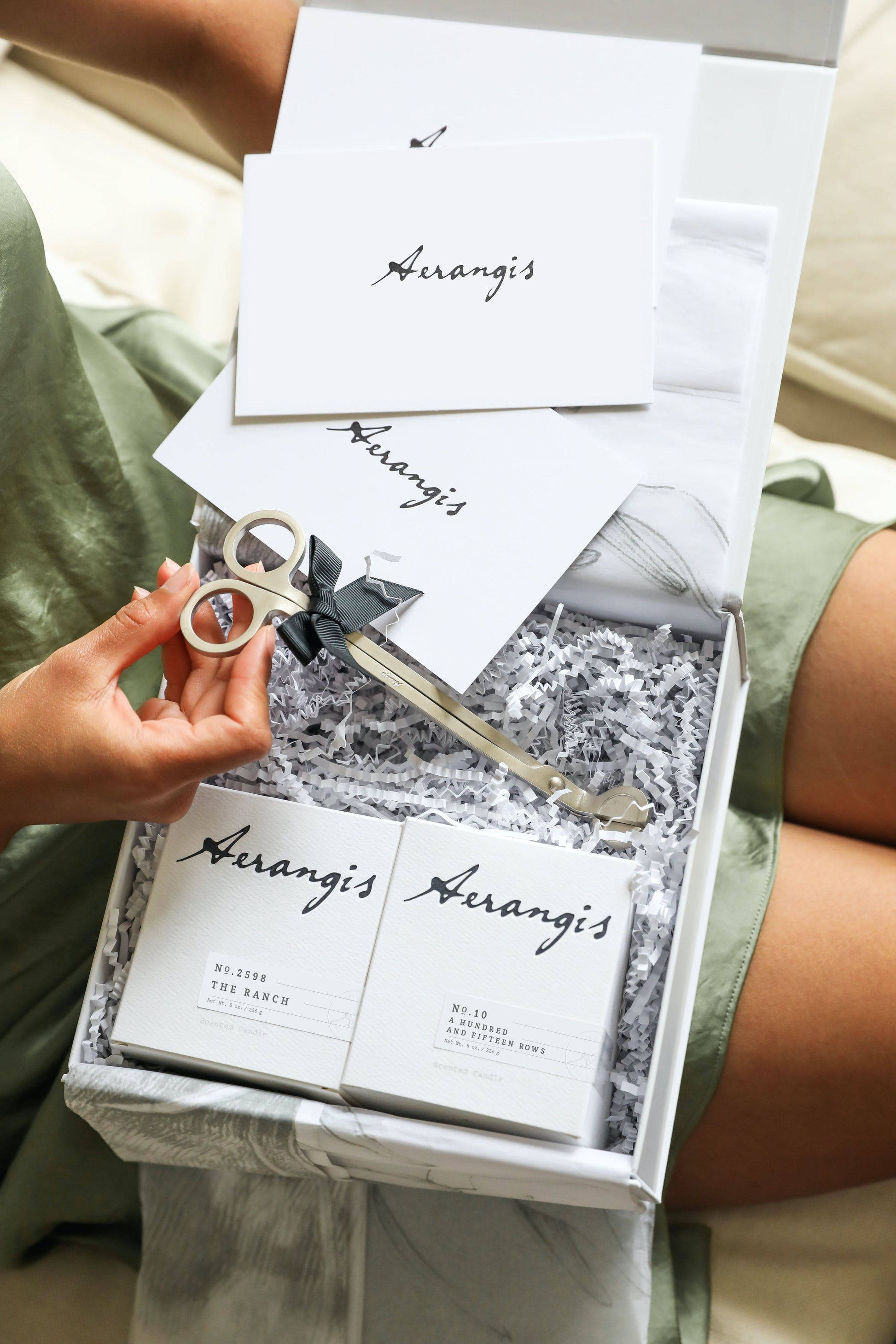 Gift box with Aerangis branded cards, two white scented candles, and silver candle wick trimmer on shredded paper fill.