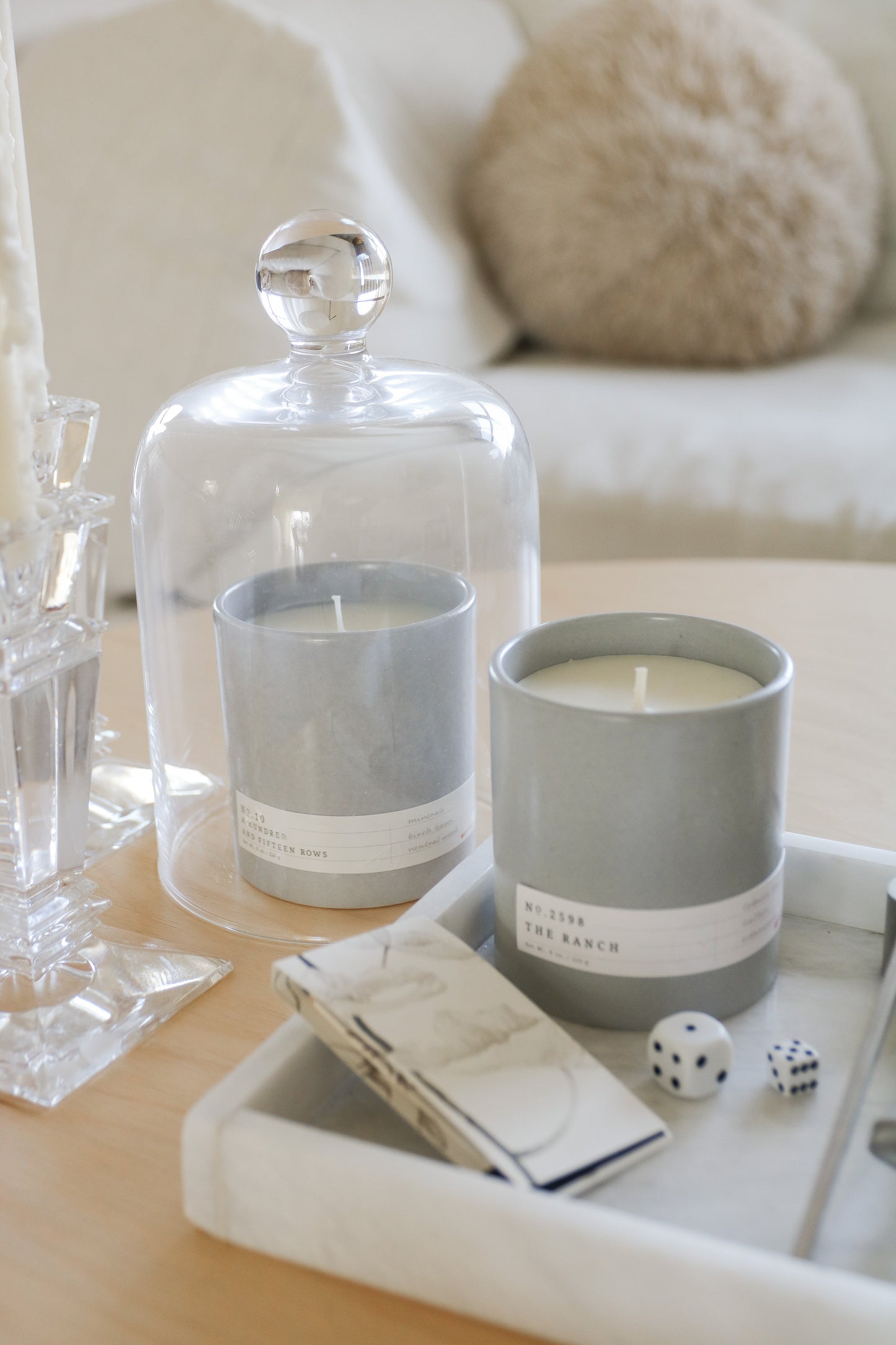 Two gray candles with white labels, one under glass cloche, on a white tray with dice and matches on wooden table