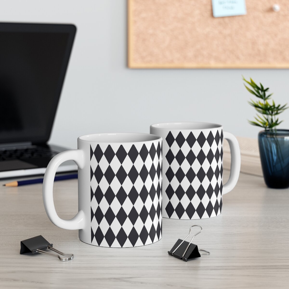 Two white ceramic mugs with black diamond pattern and large handles on a light wood desk with office supplies