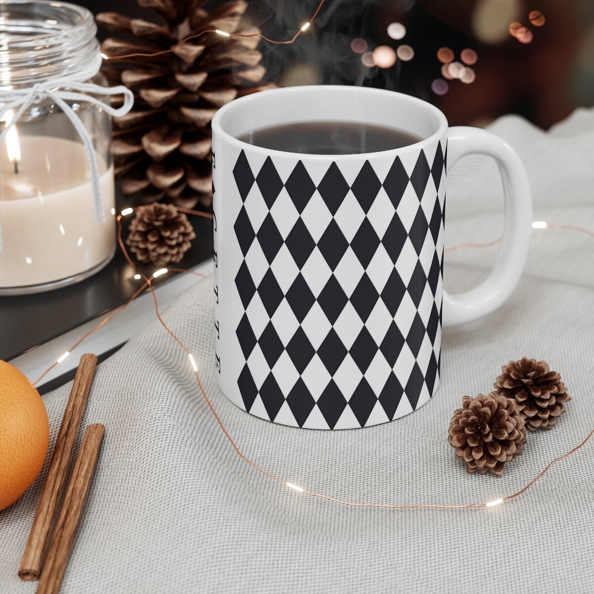 White ceramic mug with black diamond pattern filled with coffee, surrounded by pinecones, cinnamon sticks, and candlelight ambiance