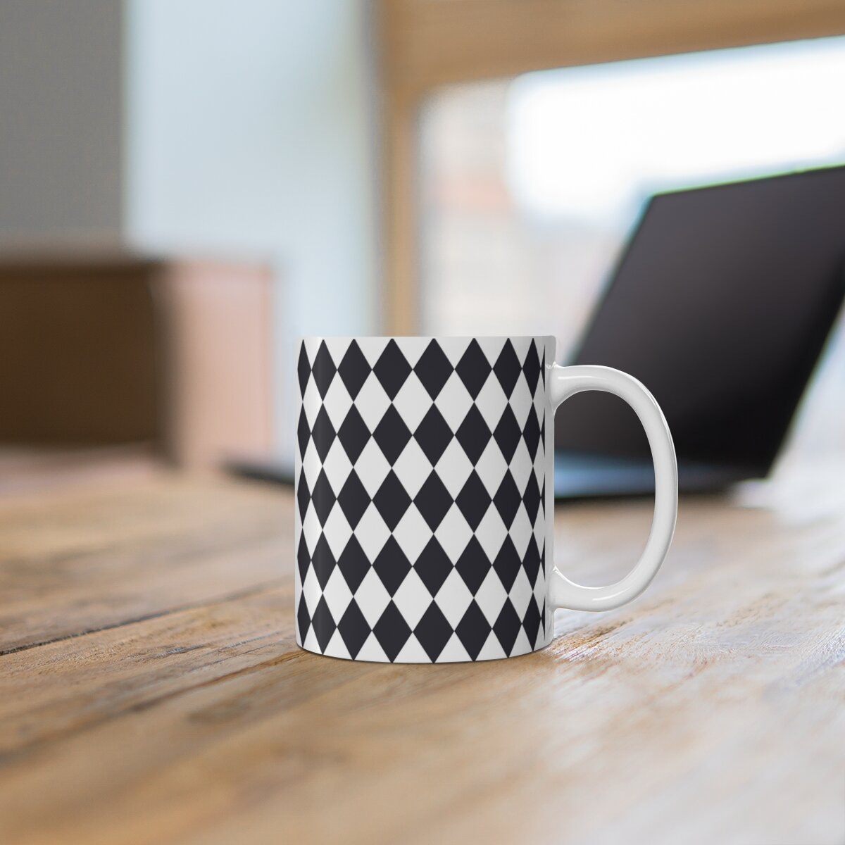 White ceramic mug with black diamond pattern and handle, placed on wooden table with blurred laptop in background